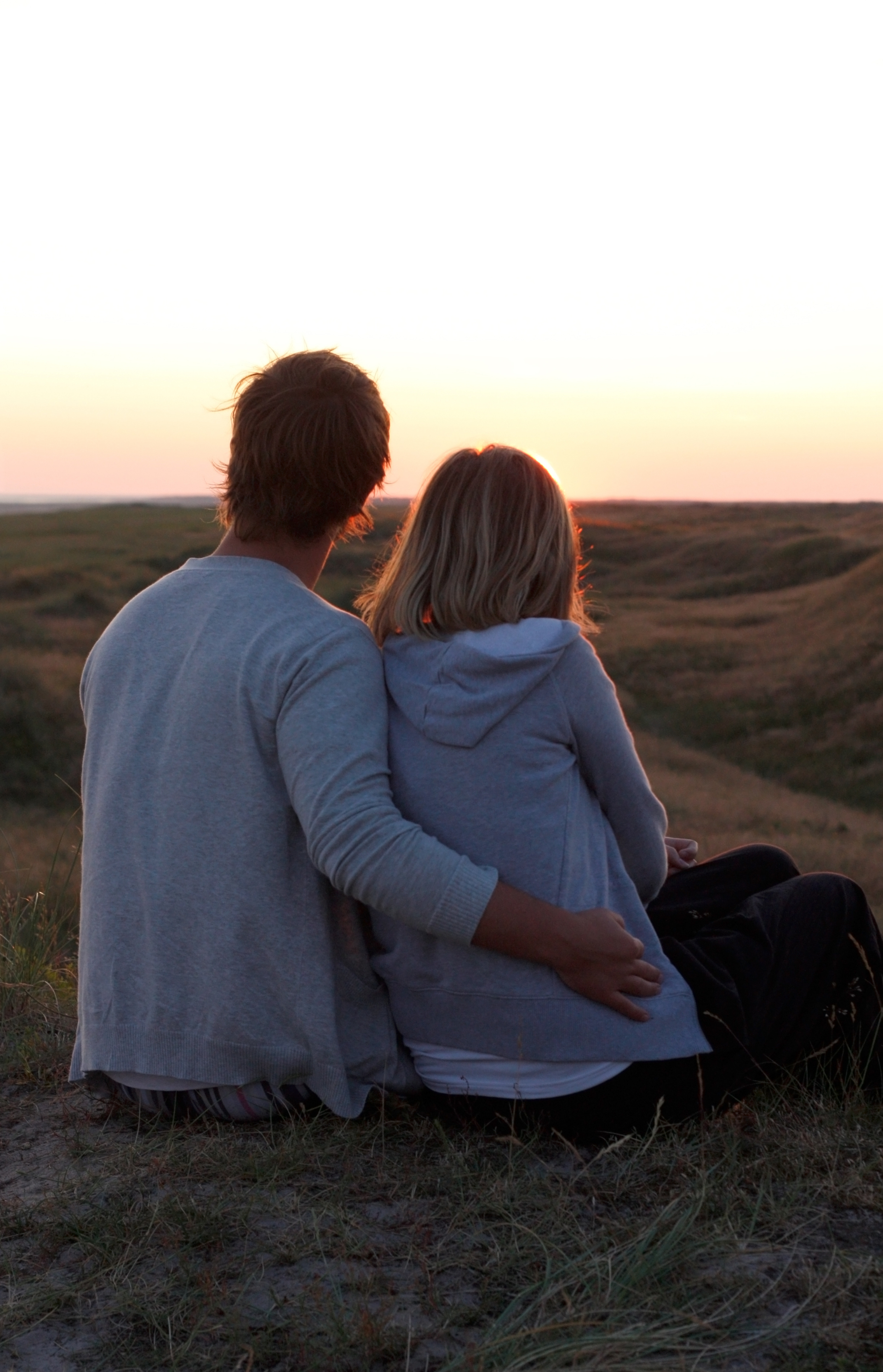 A man and a woman are sitting on top of a hill looking at the sunset.