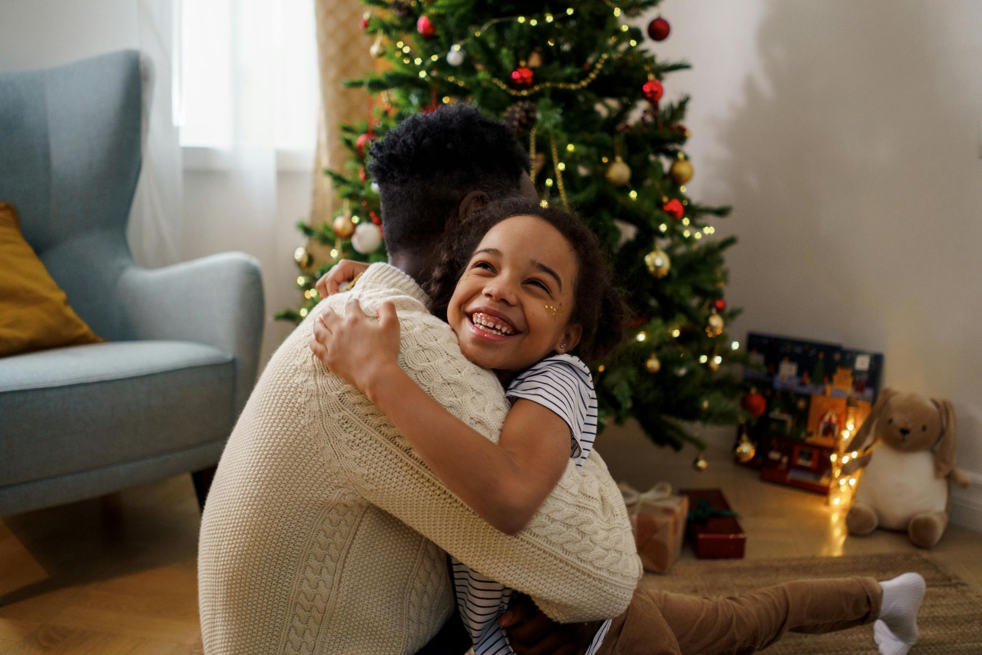 Person in a sweater hugs a smiling child in front of a decorated Christmas tree.