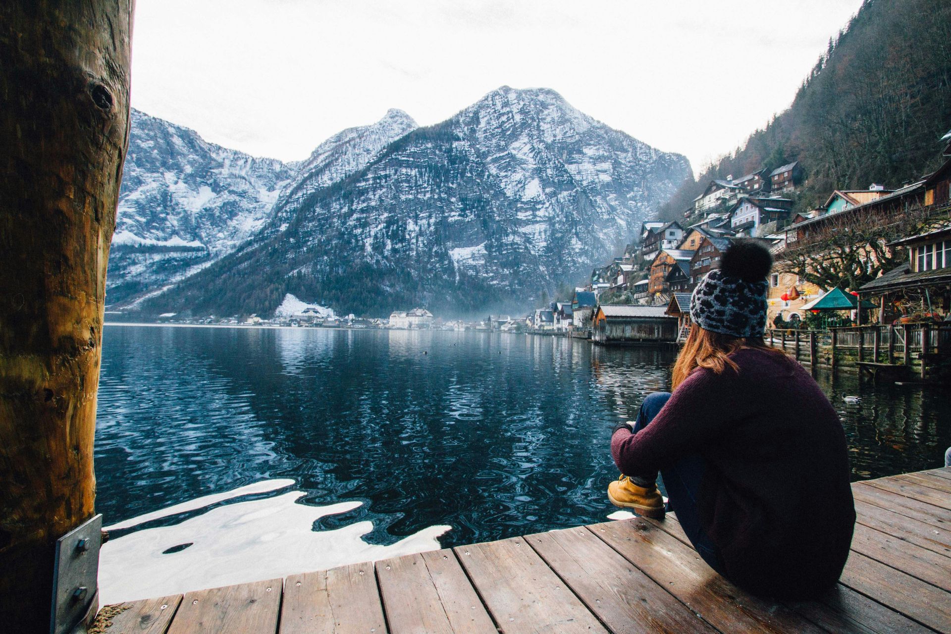 Woman sitting on a wooden dock, gazing at a snowy mountain and lakeside town.