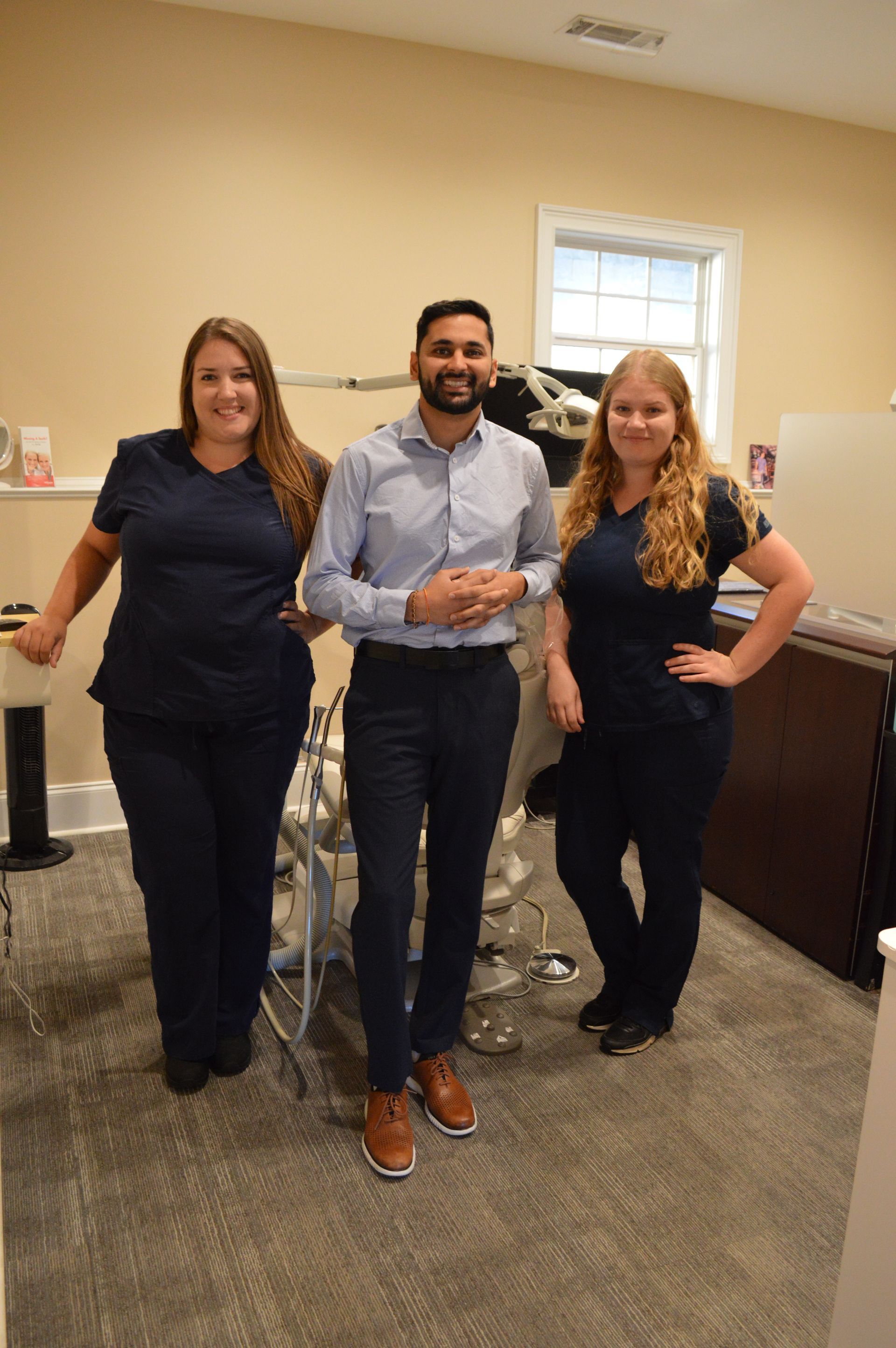 Three people standing in an office. A man in dress clothes and two women in blue scrubs smile near medical equipment.