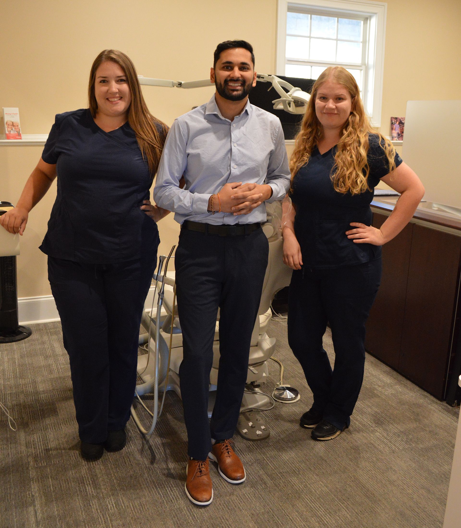 Woman with dark hair smiling, wearing teal scrubs, set against a wood-paneled wall.