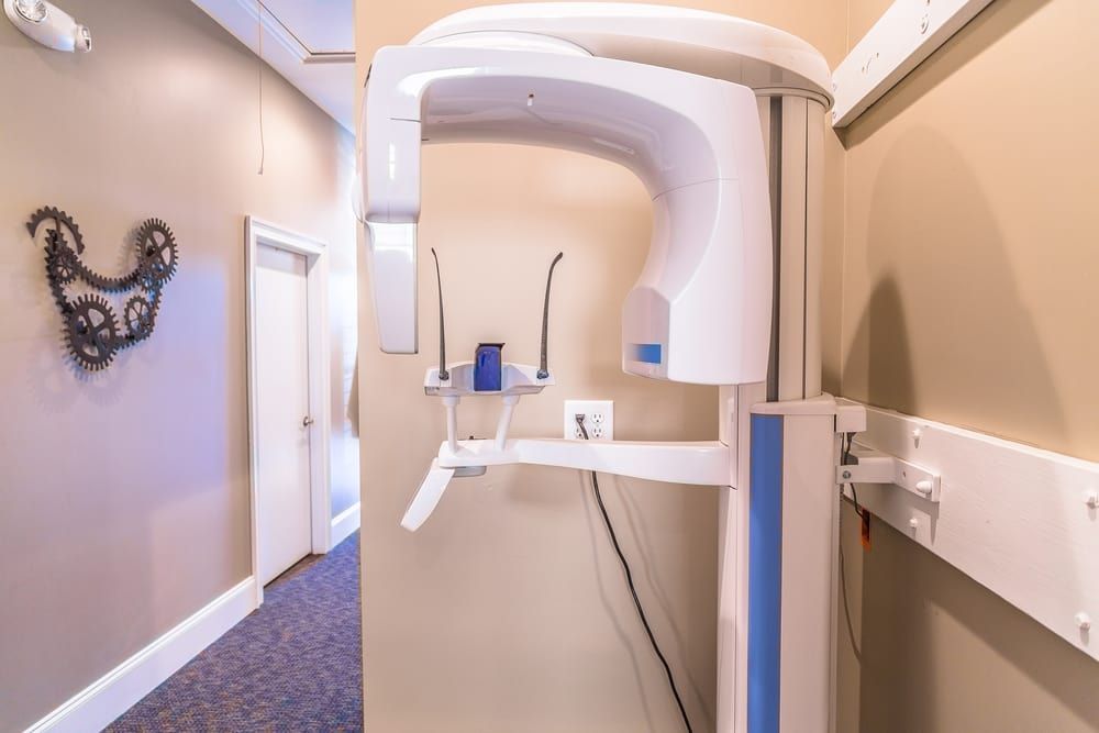 Dental X-ray machine in a beige-walled hallway. A decorative metal art piece hangs on the left wall.