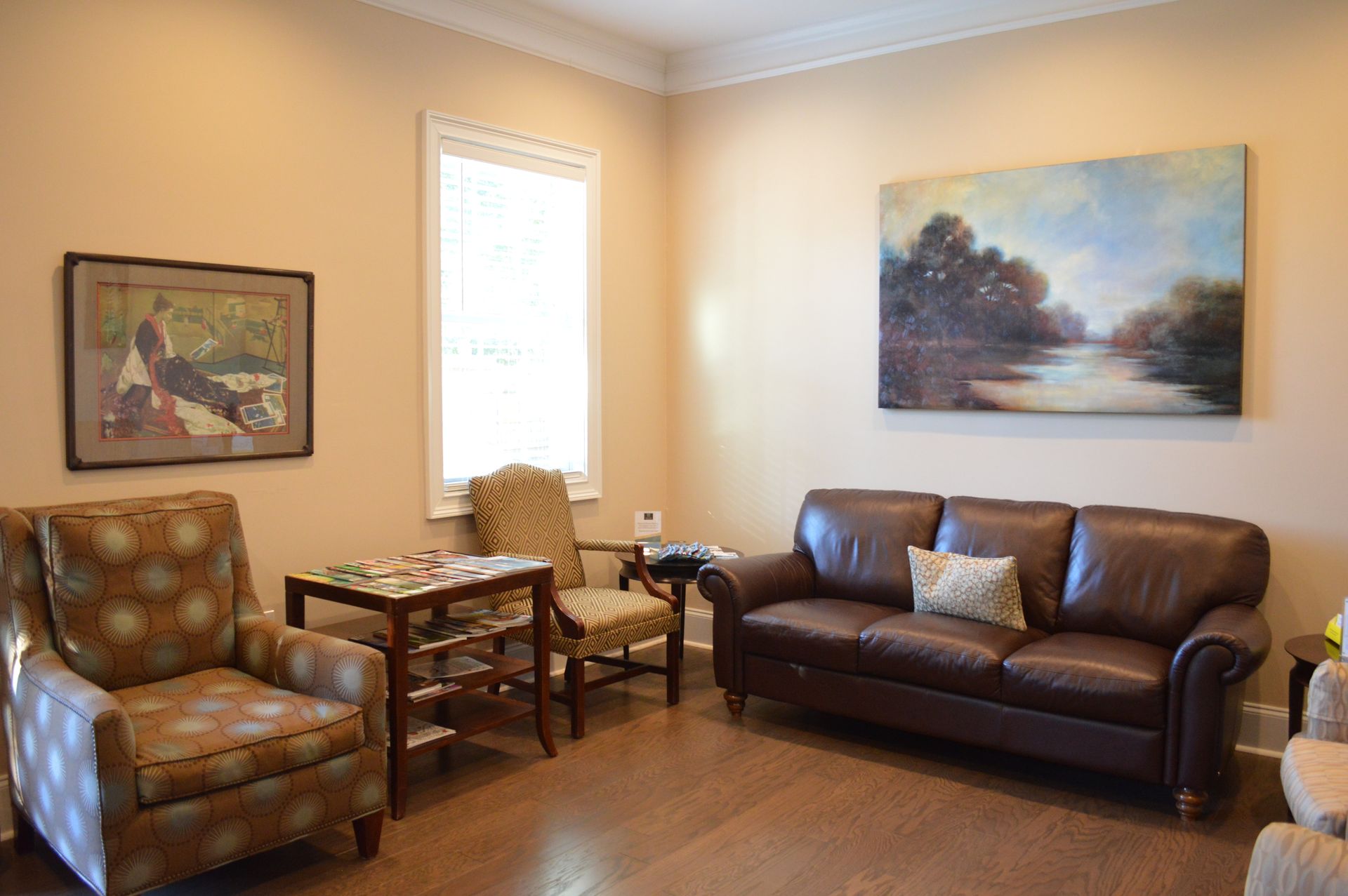 Cozy living room with brown leather sofa, patterned armchair, paintings, window, and small table.