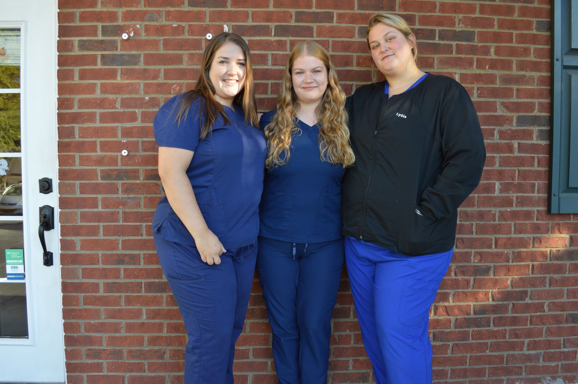 Three people in blue scrubs stand in front of a brick wall.