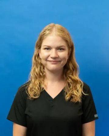 Woman with blonde wavy hair, wearing black scrubs, smiling, blue background.