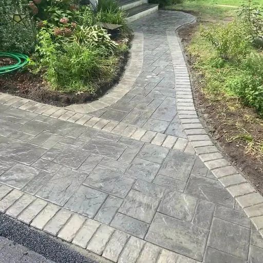 A brick walkway leading to a house with a garden in the background.