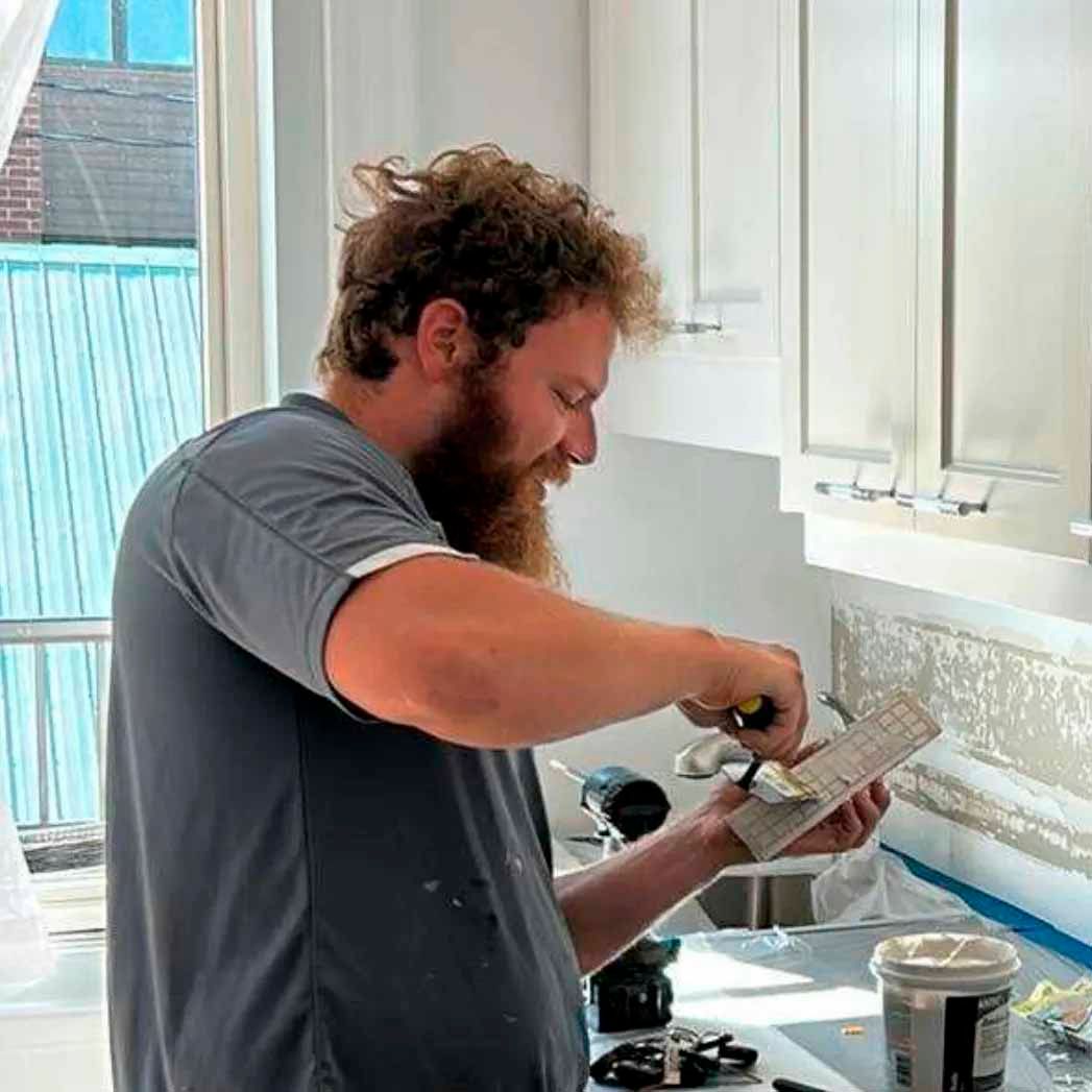Un homme pose du carrelage sur une crédence de cuisine ; il applique le joint à la truelle. Meubles et fenêtre blancs.