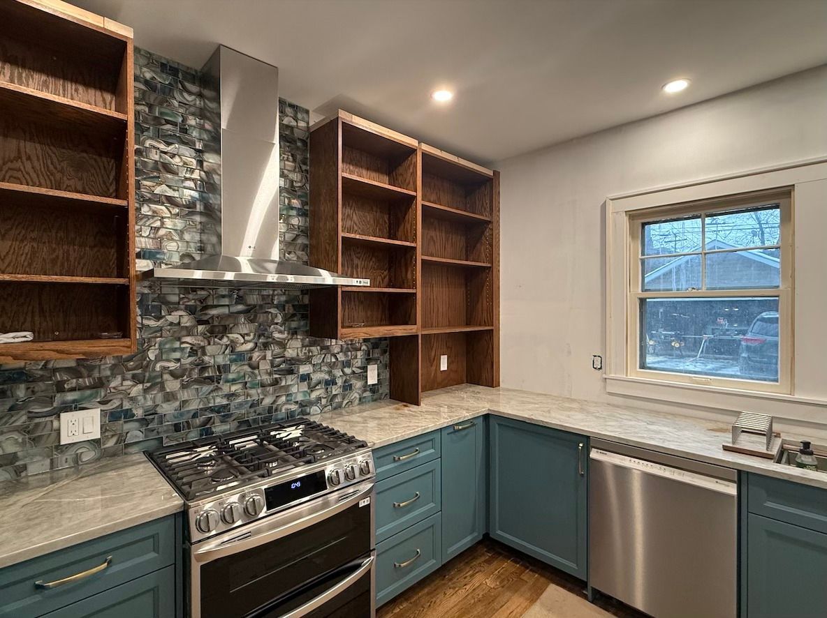 A kitchen with blue cabinets, wood shelving, a patterned mosaic backsplash, a stainless steel stove, and a window.