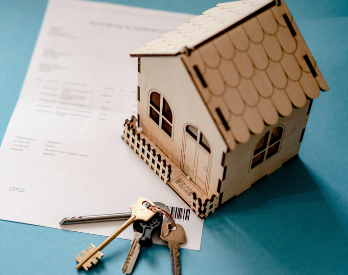 A small wooden house model and a set of metal keys rest on a document against a light blue background.