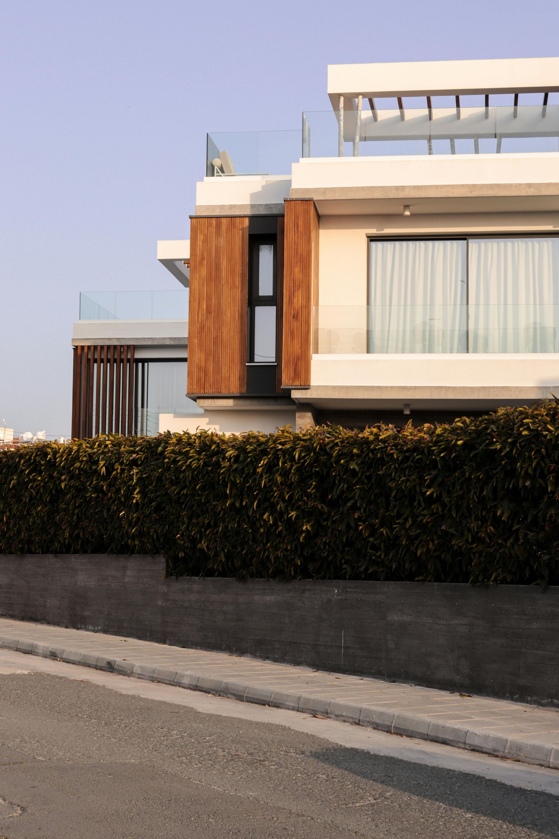Modern house with white walls, wood paneling, and a large balcony, framed by a dark stone wall and a tall green hedge.