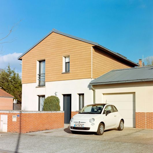 White Fiat 500 parked in front of a two-story house with tan siding and a garage. Sunny day.