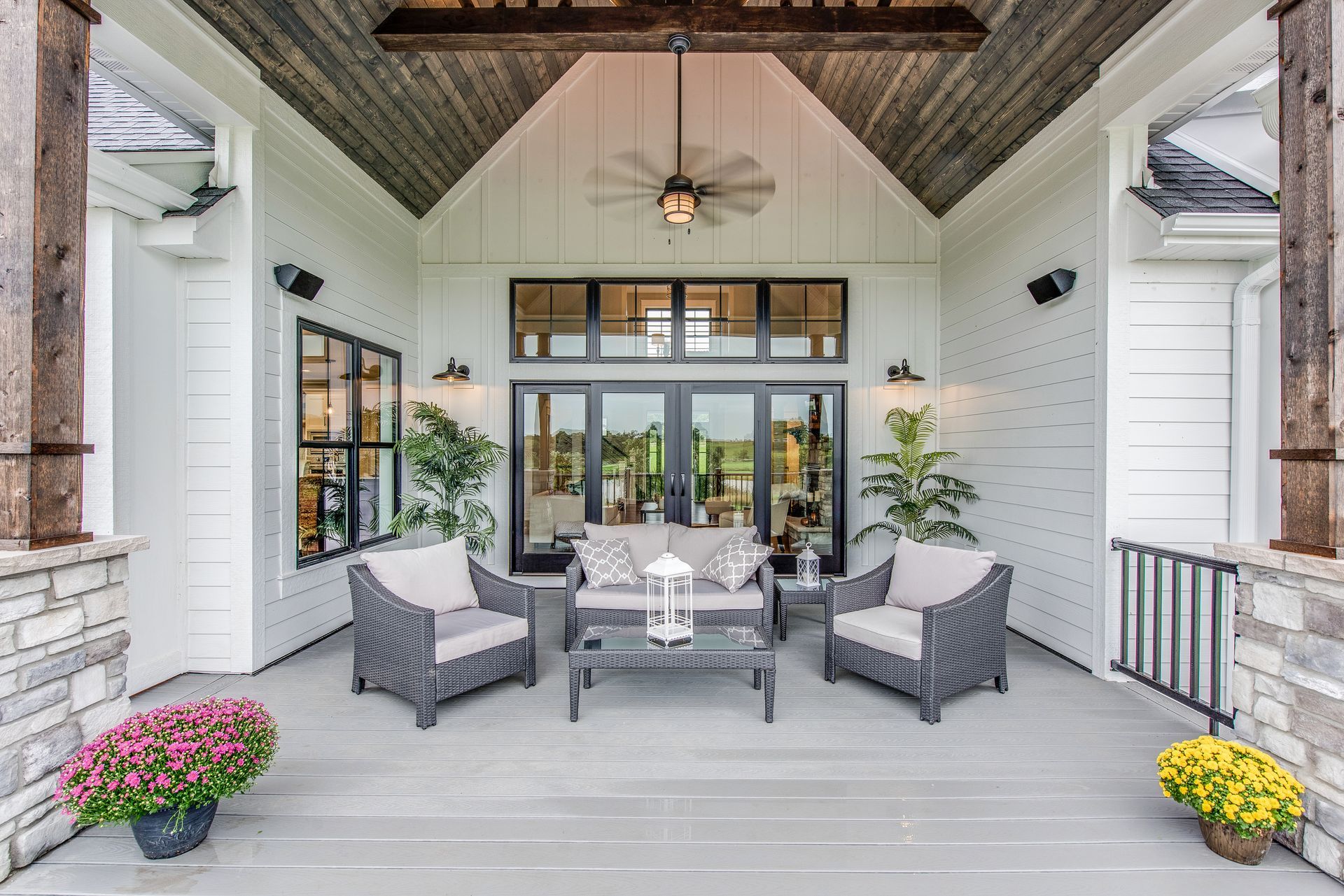 A covered porch with gray wicker furniture, a coffee table, potted flowers, and wooden accents under a vaulted ceiling.
