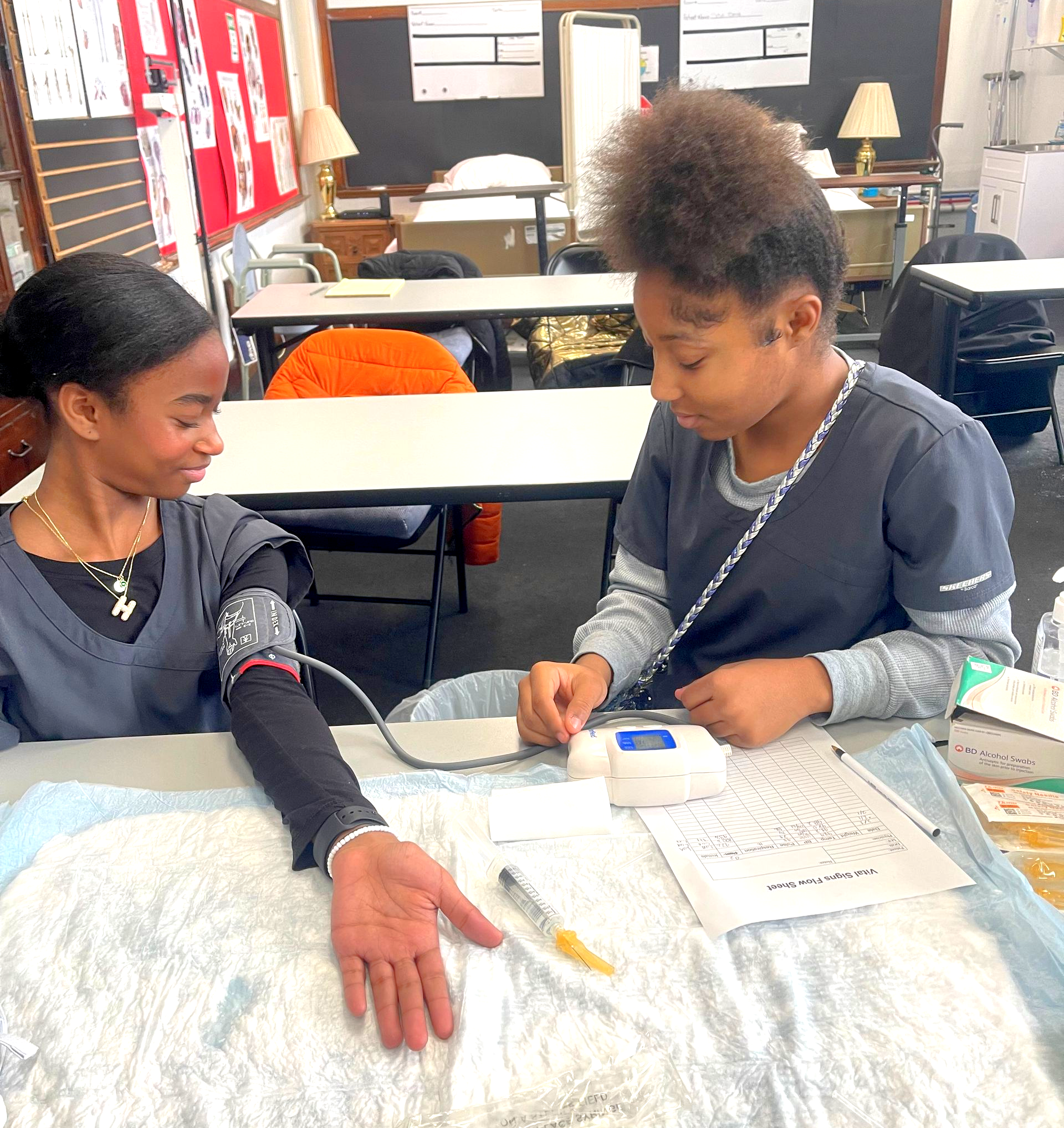 Two people in scrubs practicing blood pressure and injection techniques in a classroom.