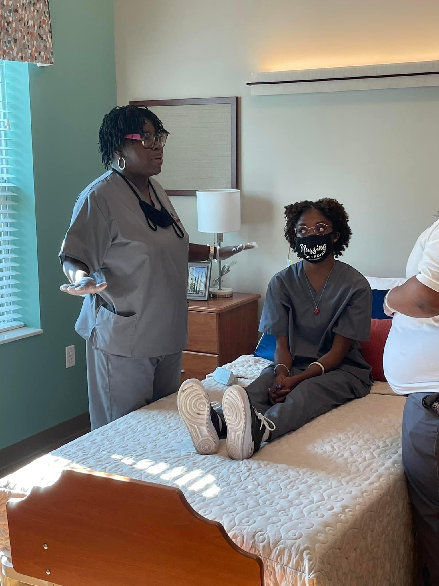 A woman in scrubs is standing next to a woman sitting on a bed.