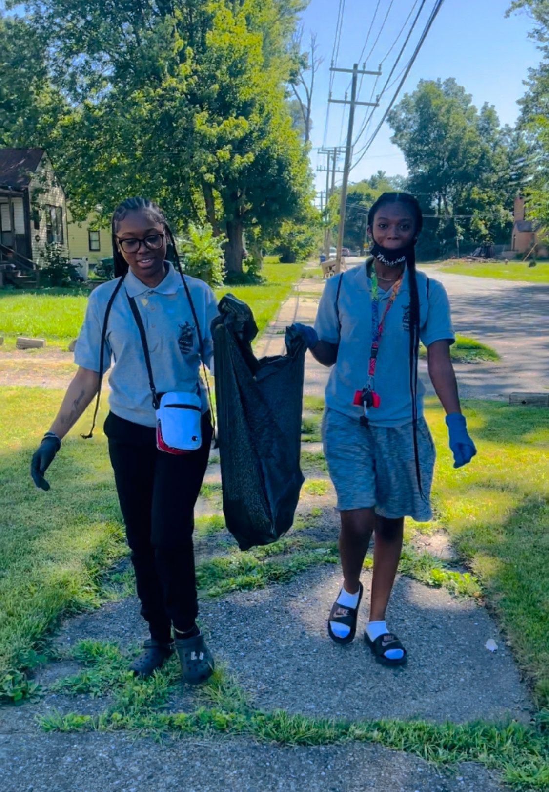 Two young women are walking down a sidewalk holding a trash bag.