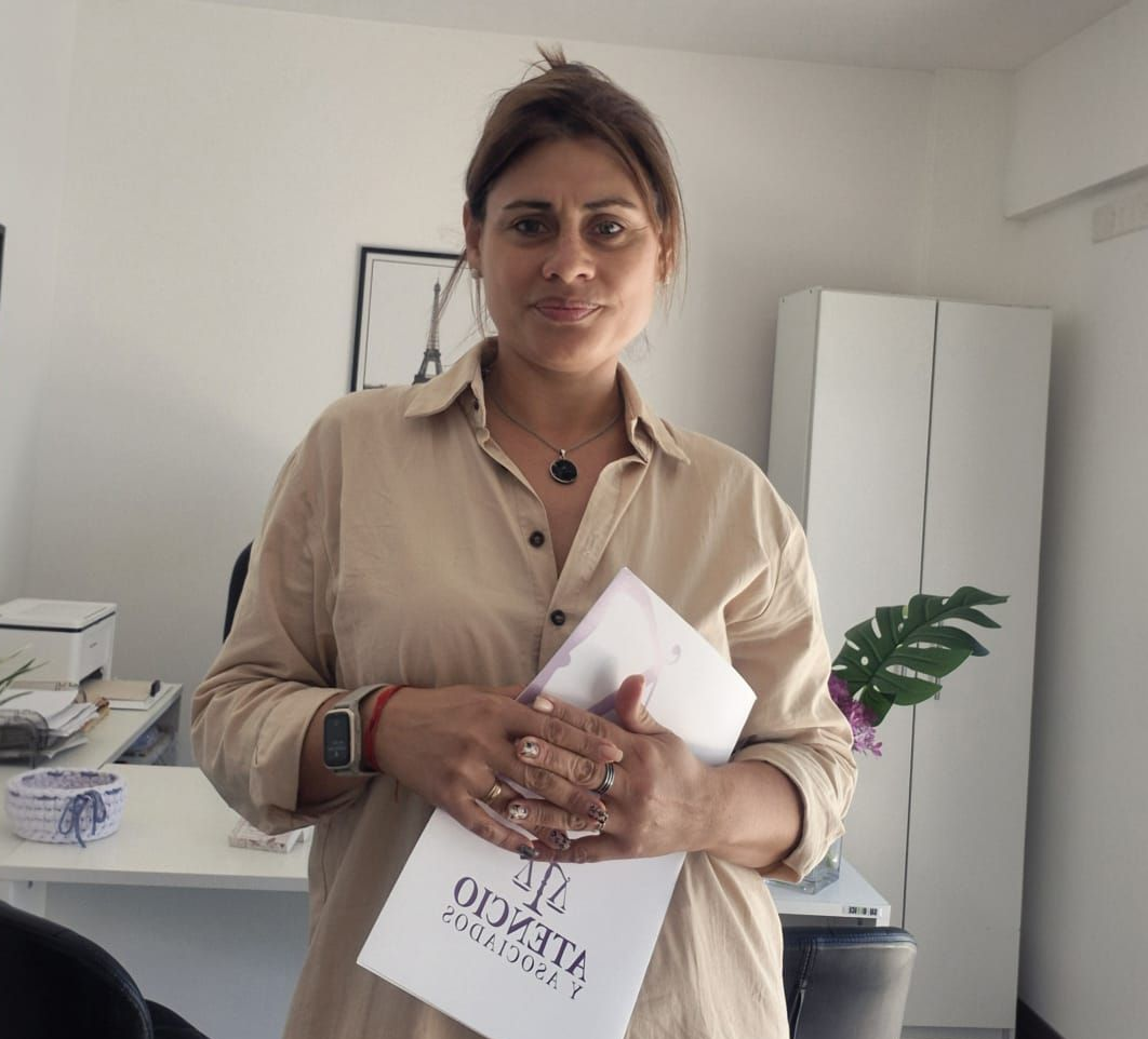 Mujer con blazer sentada en un escritorio, con las manos entrelazadas, mirando directamente al espectador,