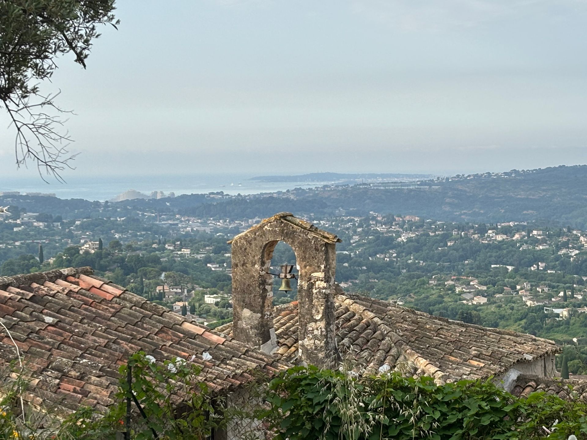 Stone bell tower with tiled roof overlooking a green valley and distant coastline under a hazy sky.