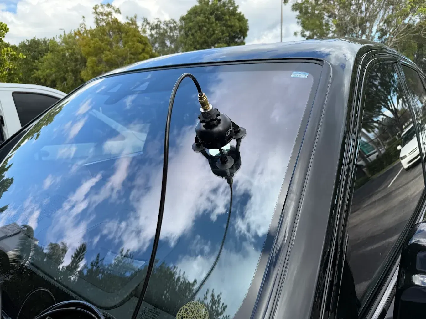 Black device attached to a car windshield, cable extending downward. Bright sky reflects on the glass.