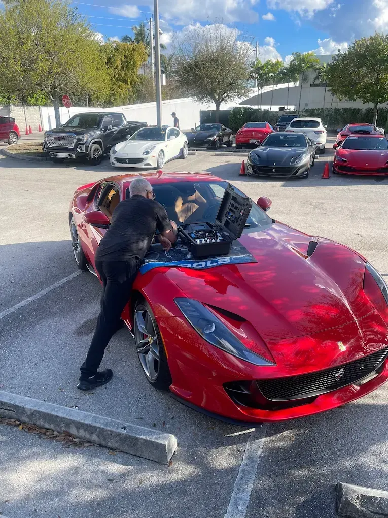 Man working on a red Ferrari in a parking lot, surrounded by other cars on a sunny day.