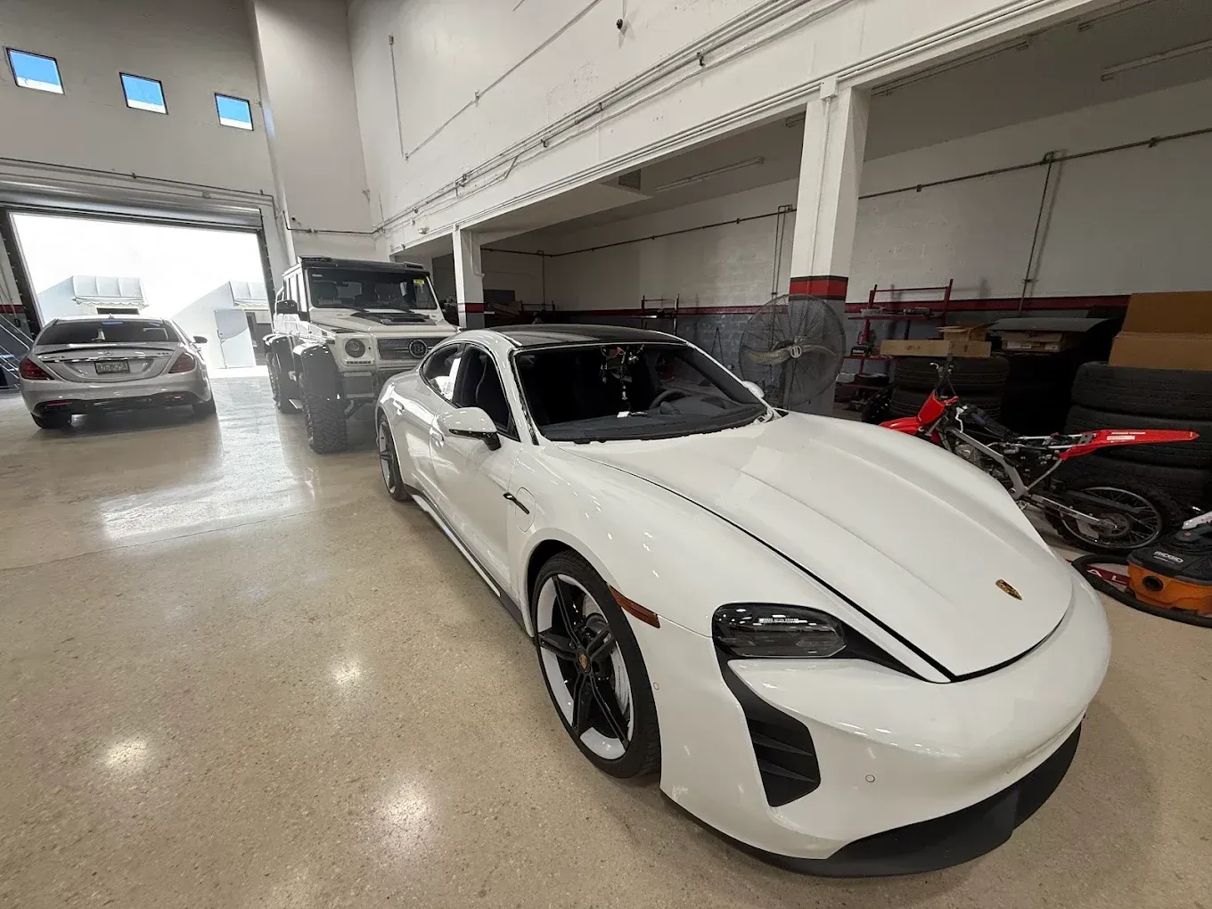 White Porsche Taycan in a garage, with a truck and car in the background.