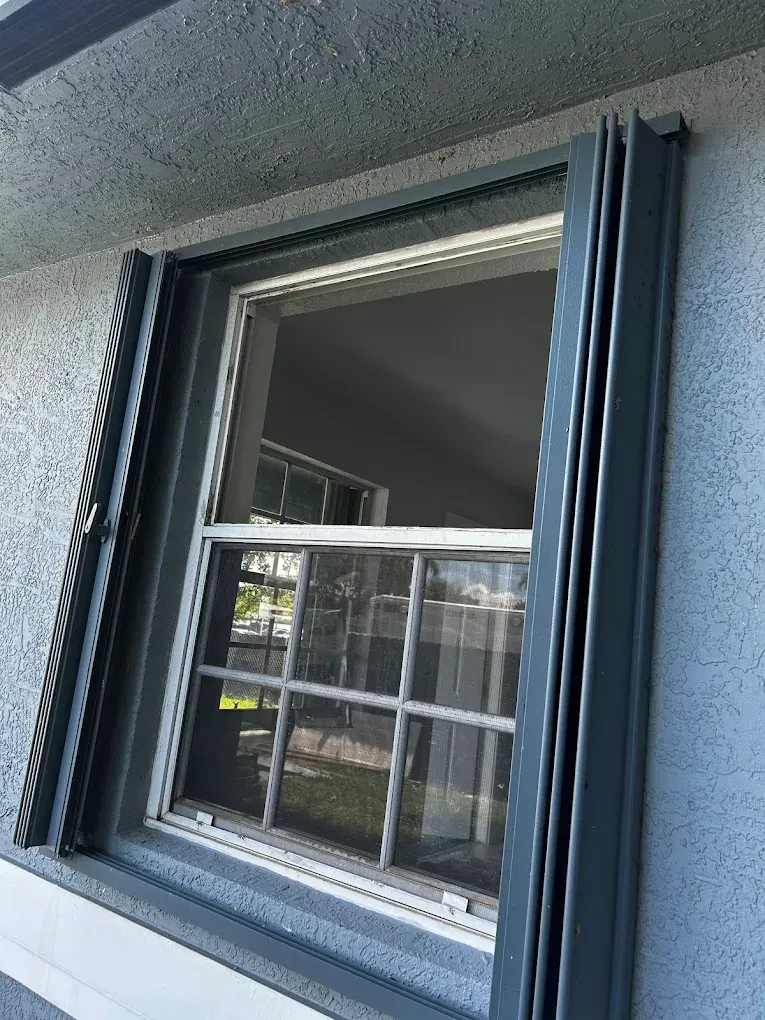 Gray-framed window with open pane, screen, and exterior shutters on a stucco wall.