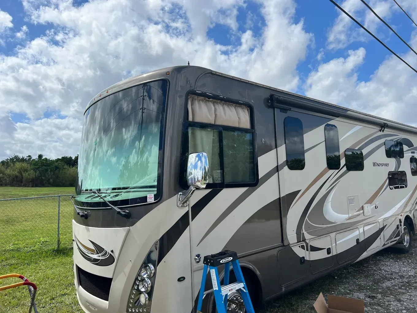 Tan and brown RV parked outdoors under a cloudy sky. A ladder is in front of the RV.