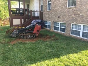 A small bulldozer is digging a hole in the grass in front of a brick house.