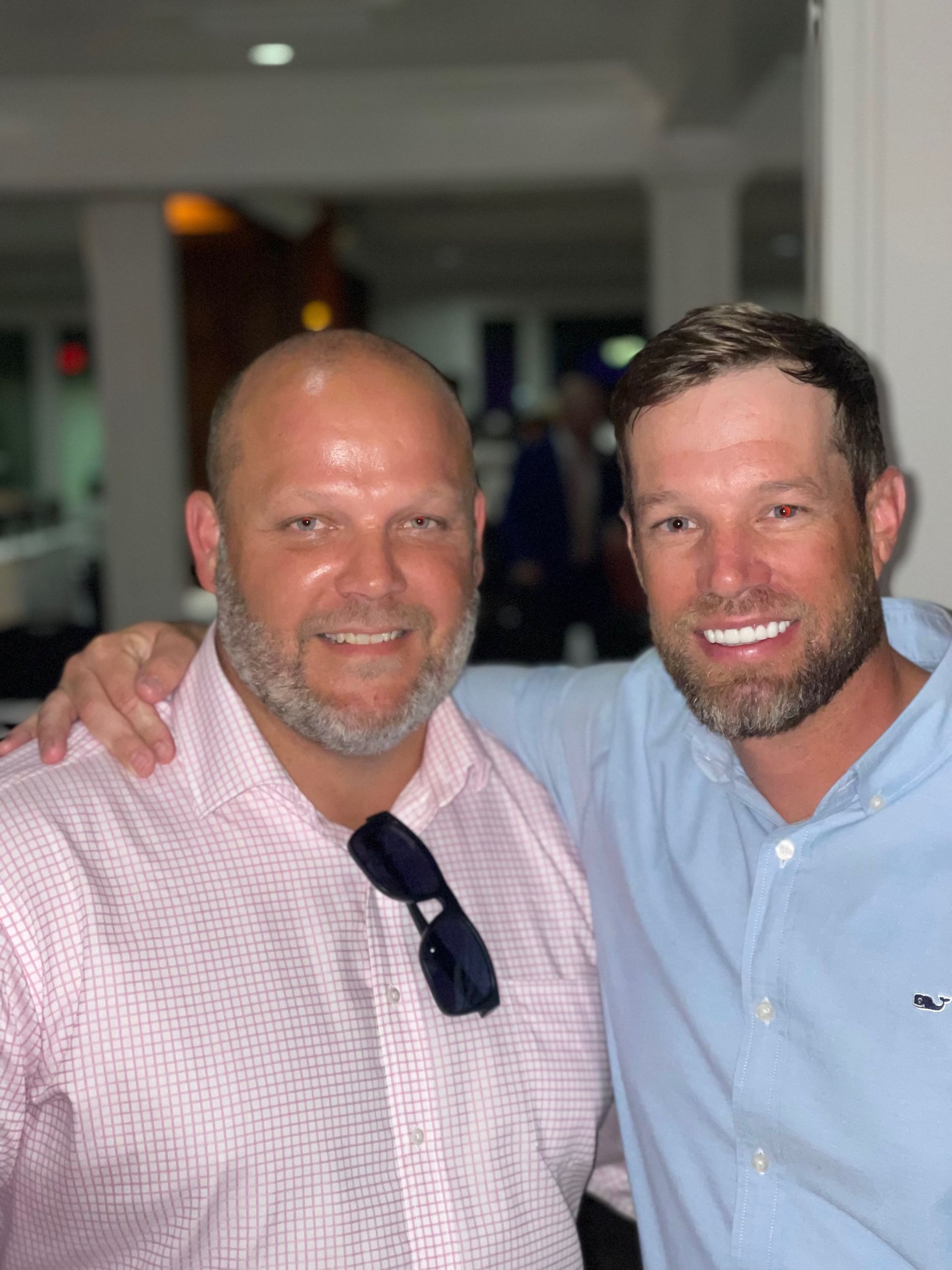 Two men smiling, shoulder to shoulder. One in a light pink shirt, the other in a blue shirt, indoors.