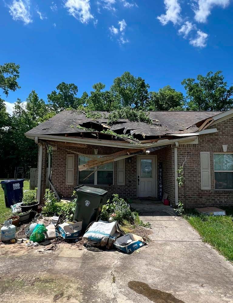 Damaged home exterior. The roof and porch are severely damaged, with debris scattered