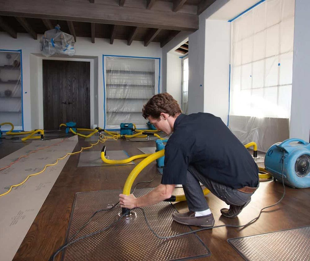 A man in a dark shirt and brown shoes kneels, attaching a hose to a floor mat in a room with drying equipment.