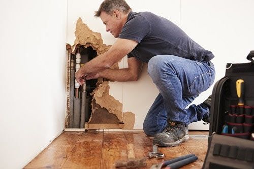 A plumber kneels, repairing pipes exposed by damaged wall, tools nearby, wooden floor, inside.
