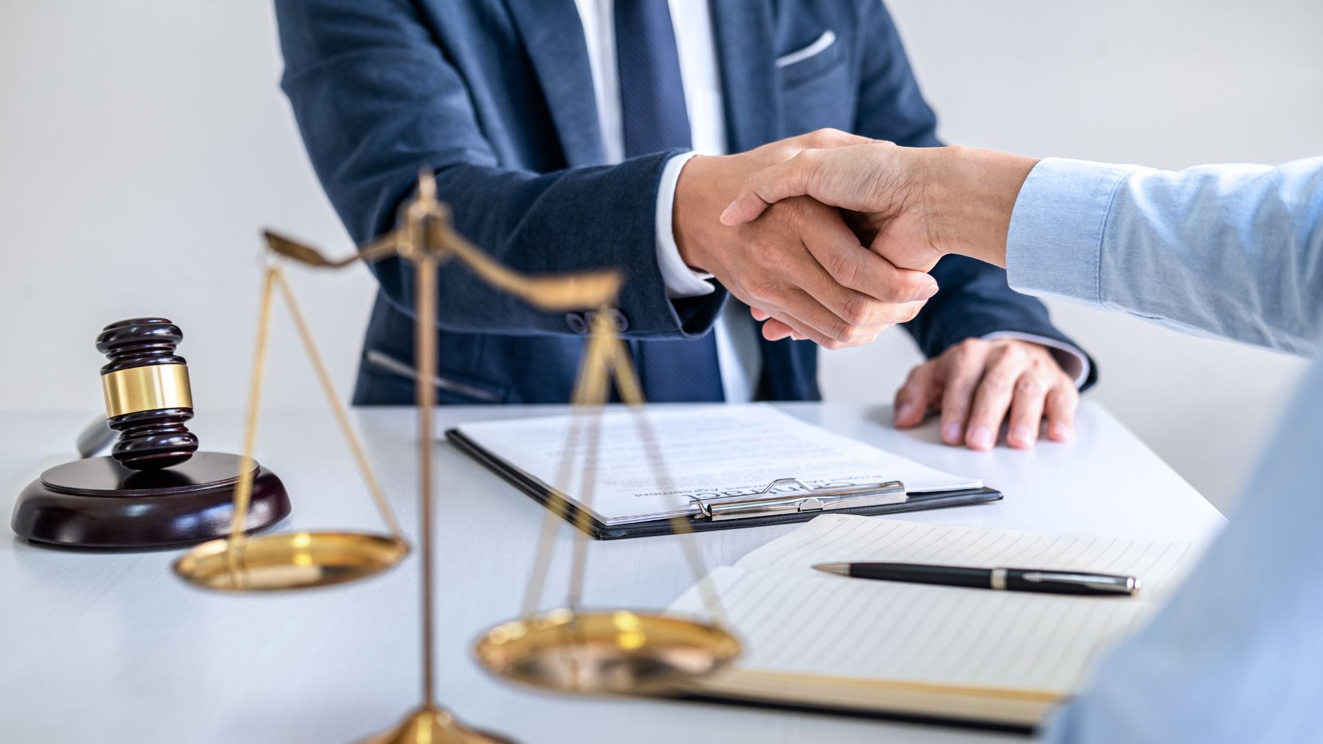 A legal professional in a suit shakes hands with a client across a desk featuring a gavel, a scale, and documents.