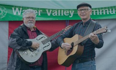 Two people stand in front of a green and red tent banner, each playing an acoustic guitar and smiling at the camera.