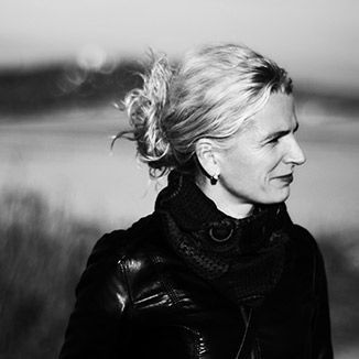 A woman in a black jacket and scarf is standing in front of a body of water.