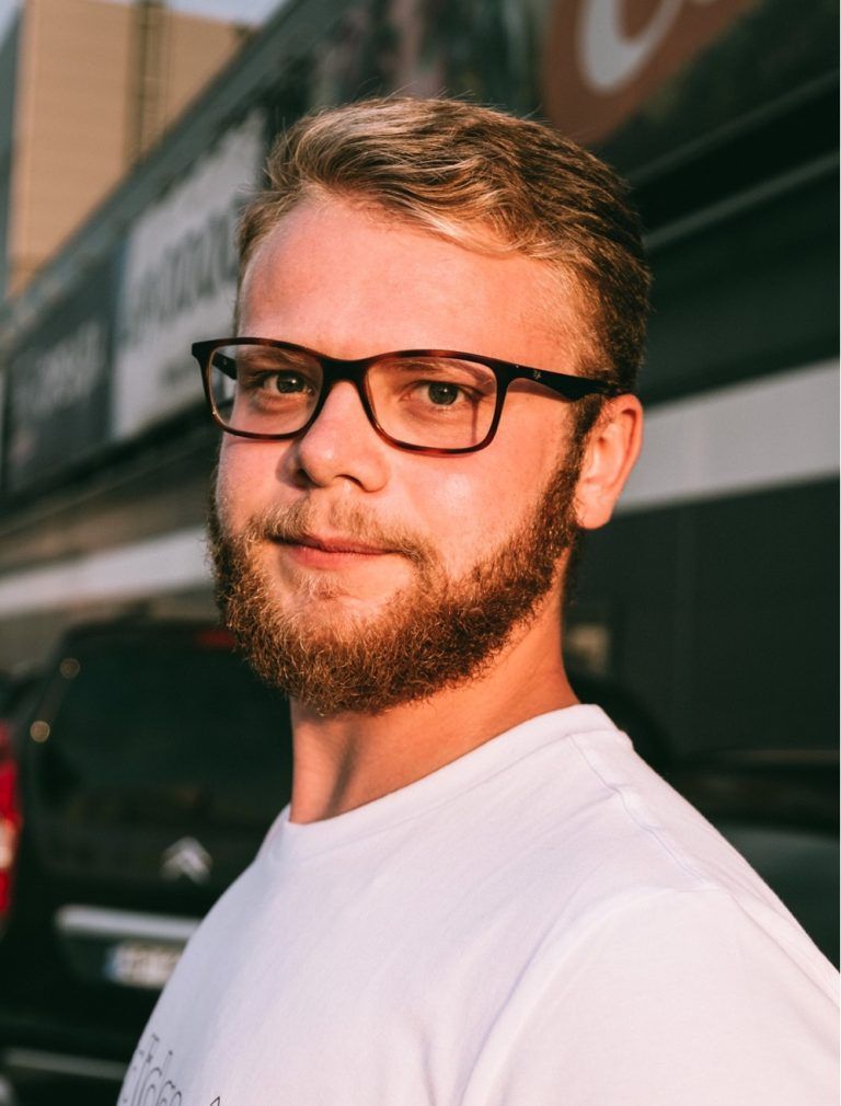 A man with a beard and glasses is standing in front of a building.