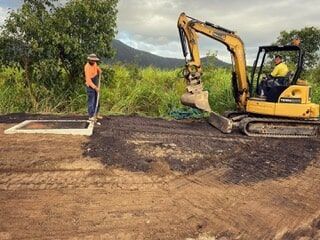 A Man Is Standing Next to A Yellow Excavator in A Dirt Field — Terranovus Earthworks & Plant Hire in Cairns, QLD