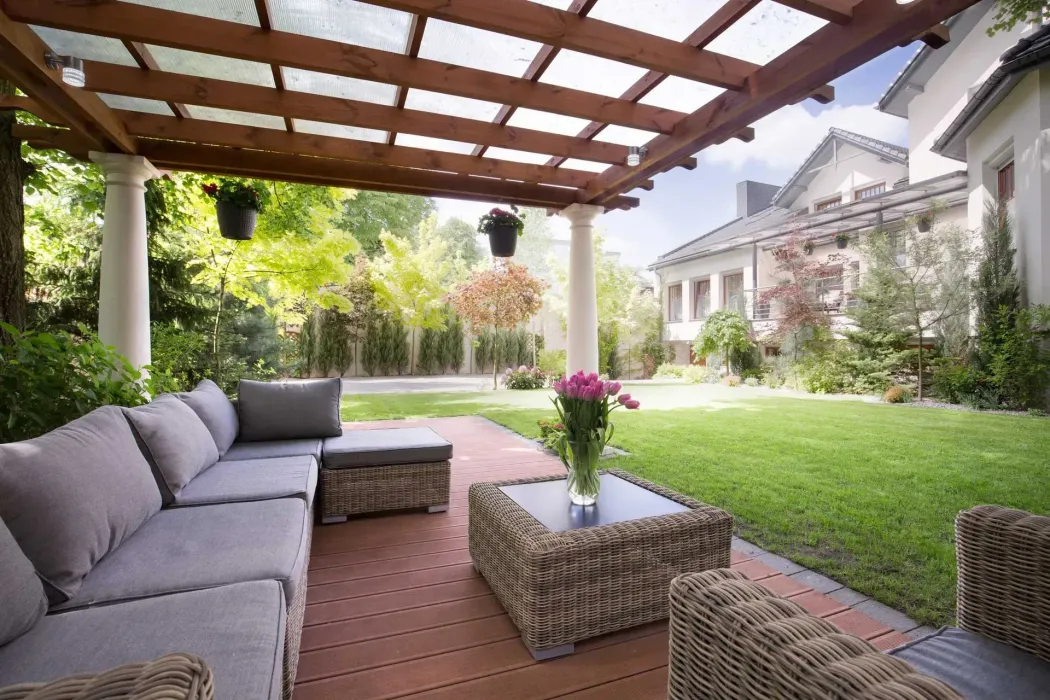 Outdoor patio with wicker furniture under a wooden pergola, facing a manicured lawn and house. Outdoor patio with wicker furniture under a wooden pergola, facing a manicured lawn and house.