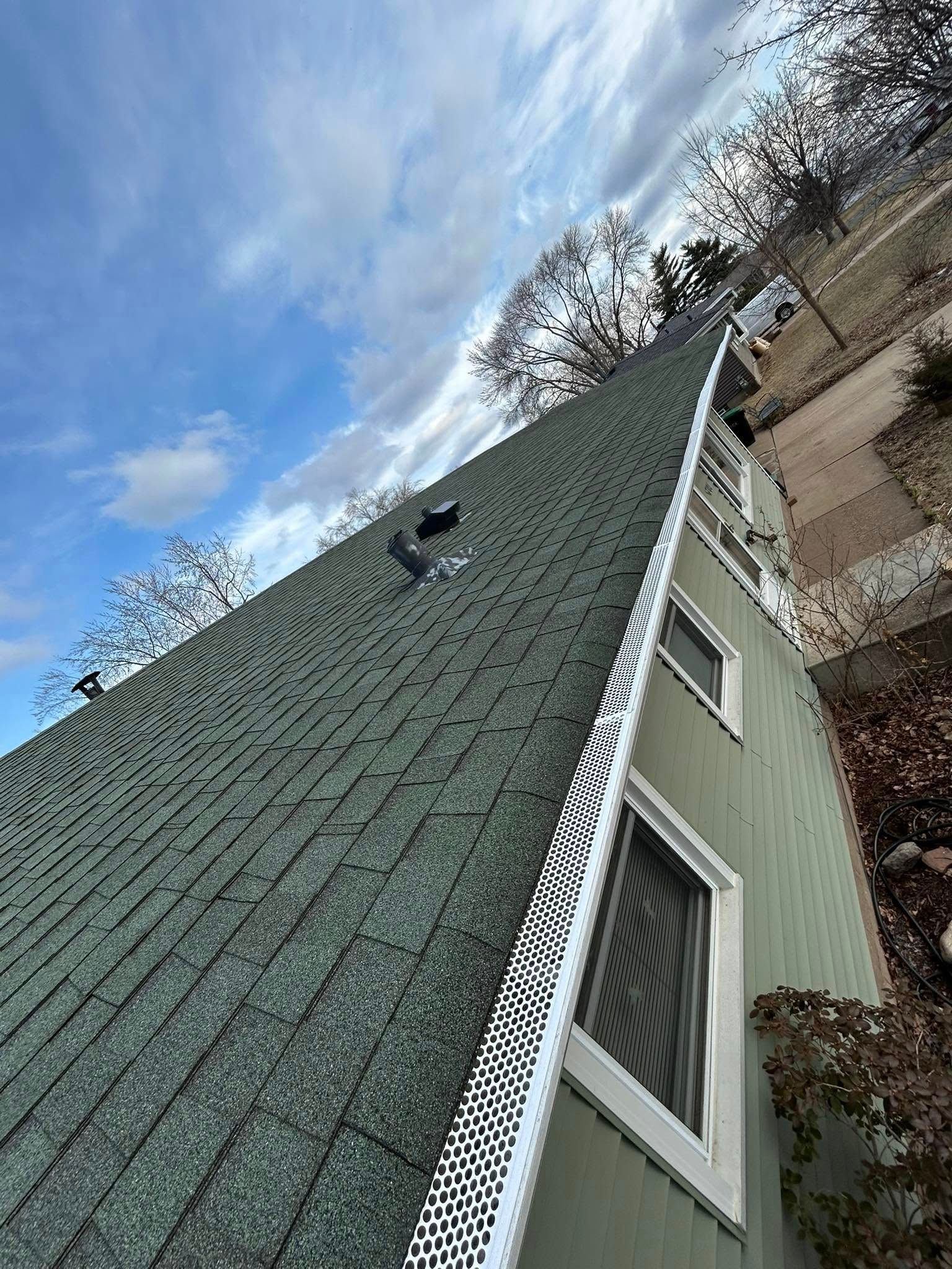 Green shingled roof of a house with white trim. Blue sky with clouds.