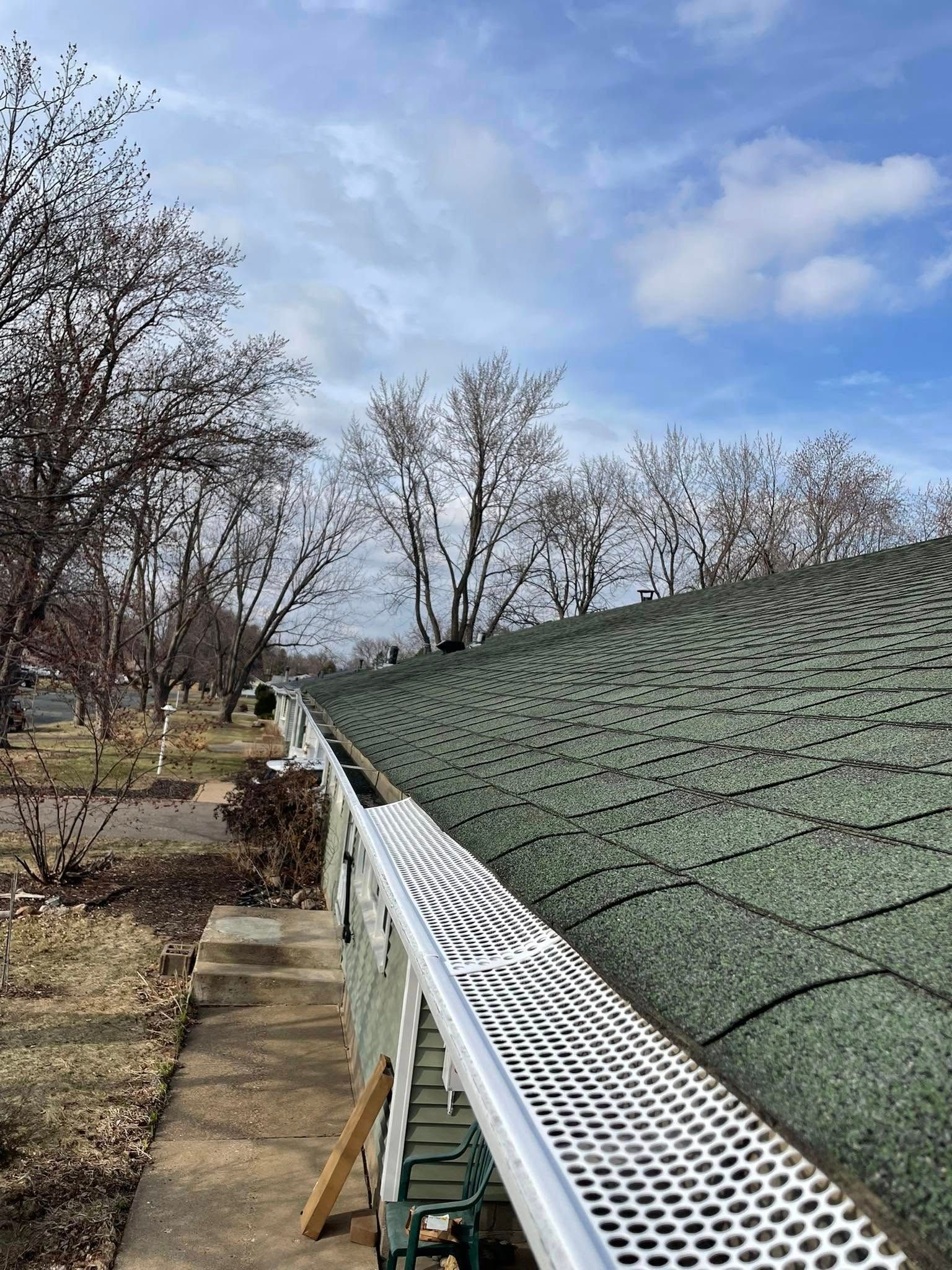 Gutters on a house with perforated covers. Green roof, trees, and cloudy sky.