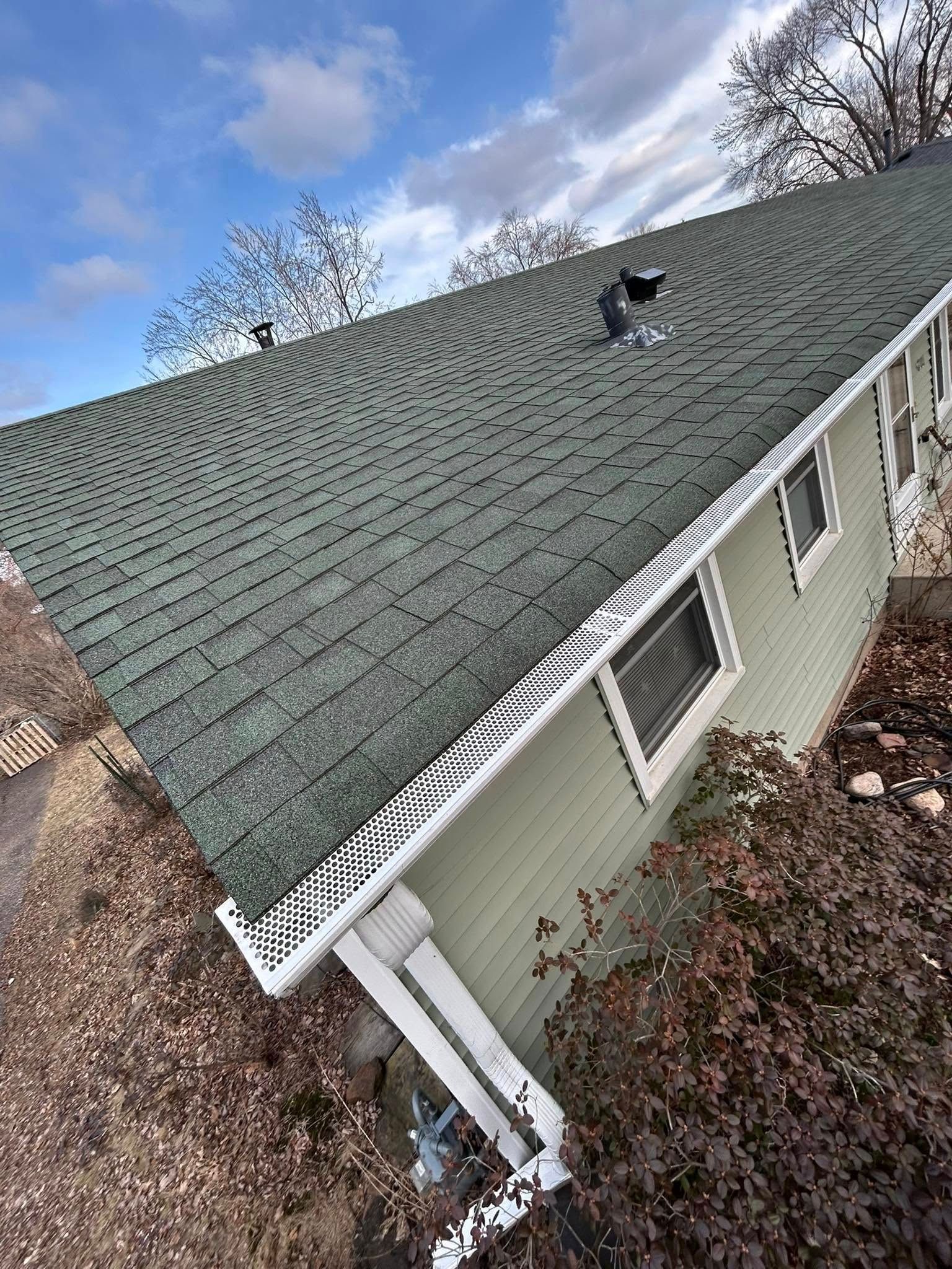 Green shingled roof of a house with white trim against a cloudy blue sky.