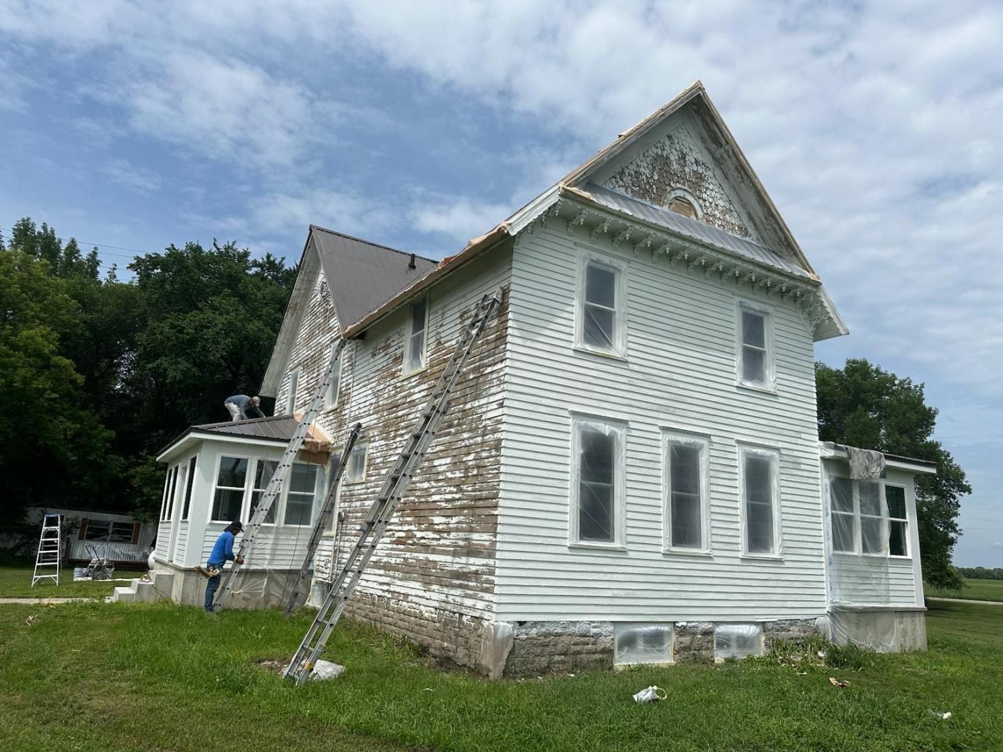 Two-story white house with peeling paint, person on ladder; partly cloudy sky, green grass.