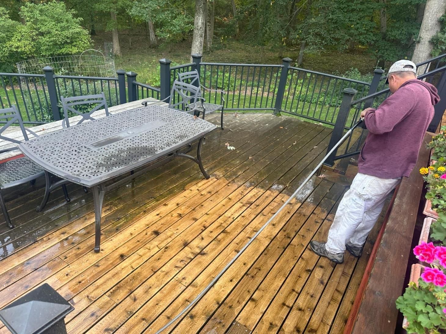 Person power washing a wooden deck with a metal table and chairs, surrounded by railing and greenery.