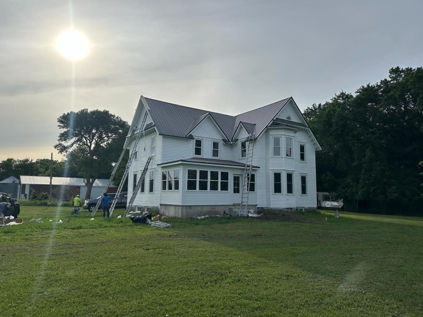 White farmhouse with new gray metal roof, surrounded by green grass; workers with ladder.