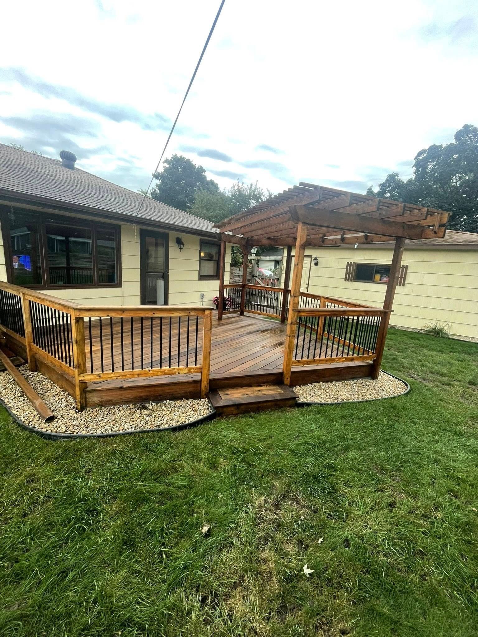 Wooden deck with pergola in backyard, beige house backdrop, gravel border, green grass, cloudy sky.
