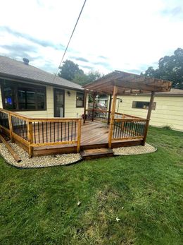 Wooden deck with pergola in backyard, beige house backdrop, gravel border, green grass, cloudy sky.