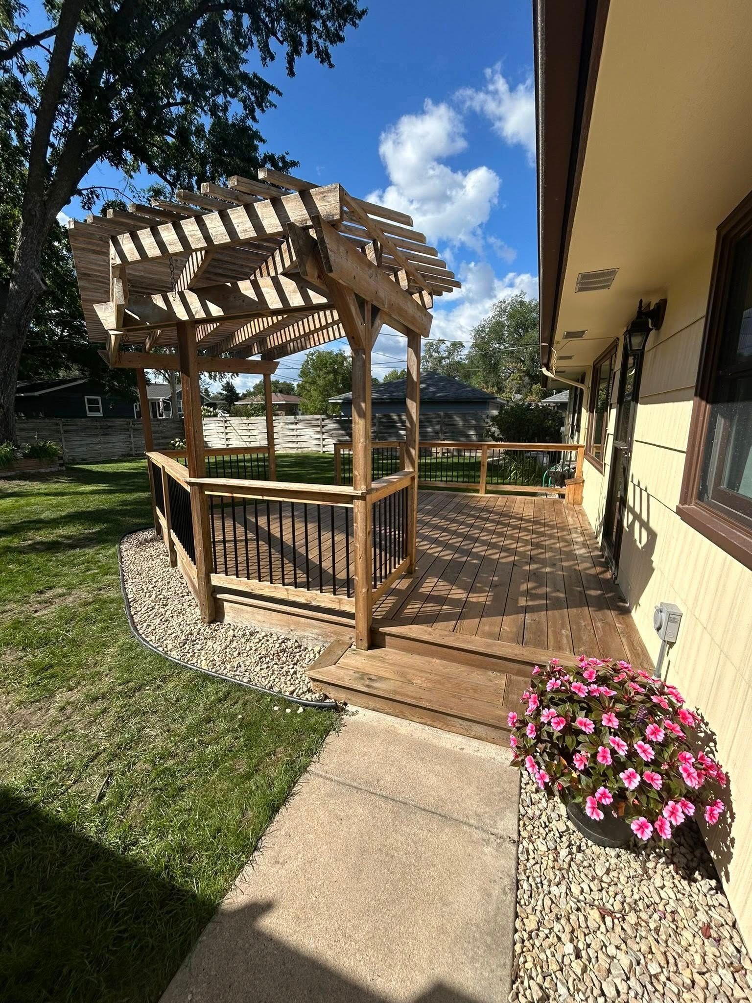 Wooden deck with pergola, surrounded by rock, beside a tan house with pink flowers.