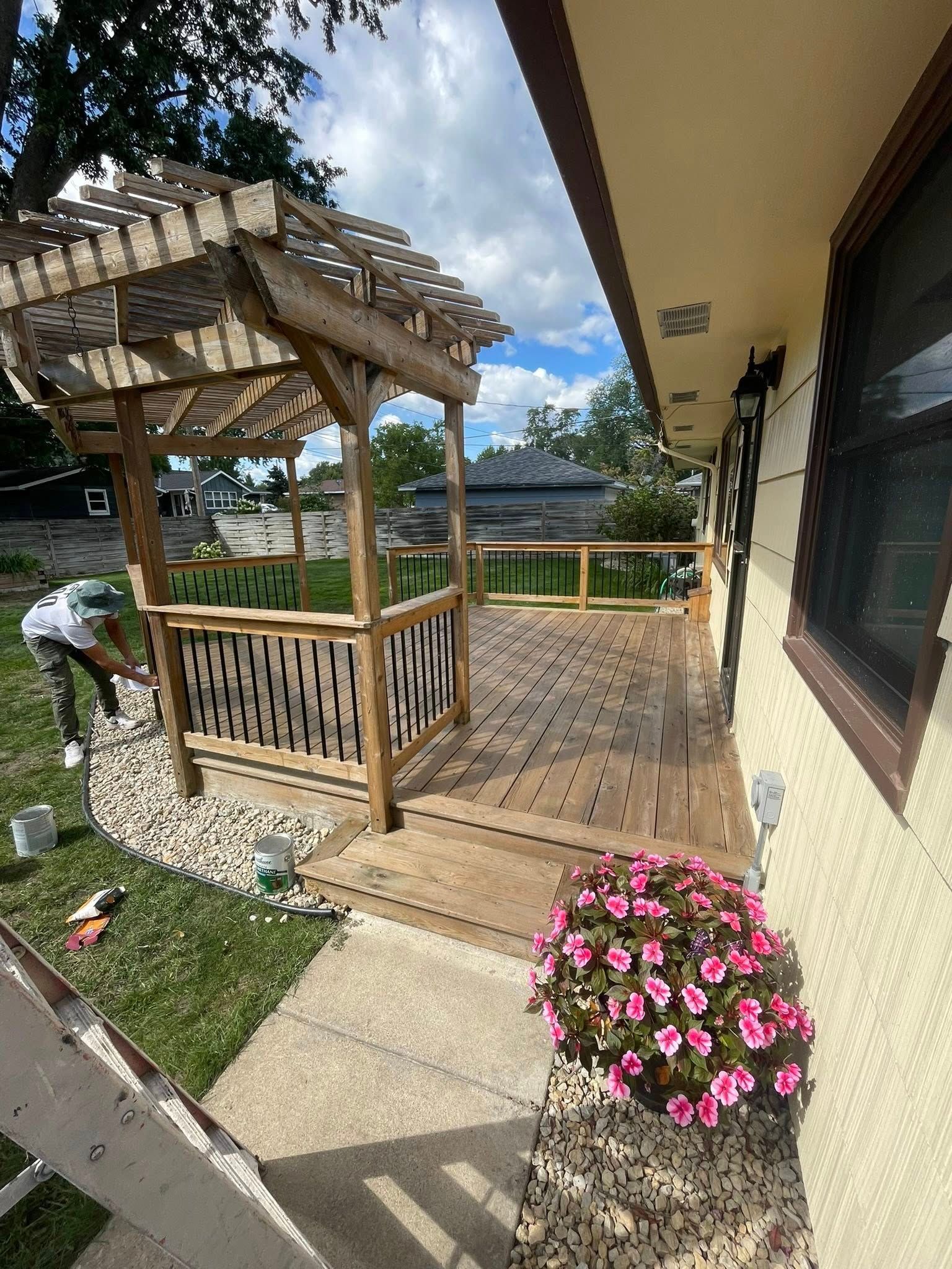 Wooden pergola and deck on a beige building exterior with a person painting; pink flowers in foreground.