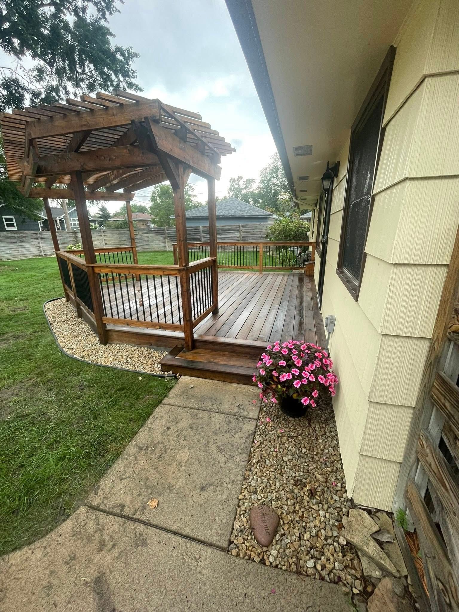 Wooden pergola on a deck beside a house with a flower pot. Gravel and grass surround it.