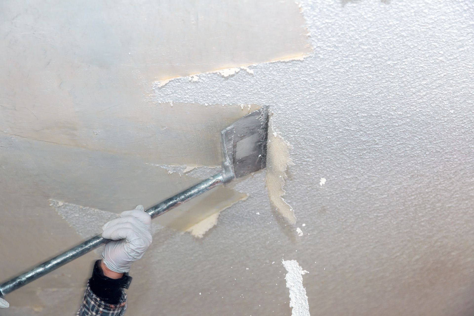 Person scraping textured ceiling with a tool, wearing gloves, in a white room.