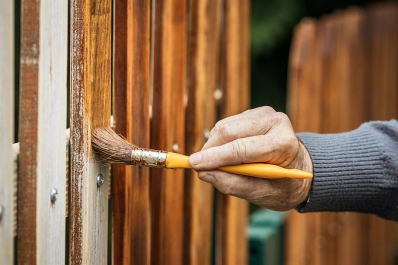 Hand painting a wooden fence with a brown brush and stain outdoors.